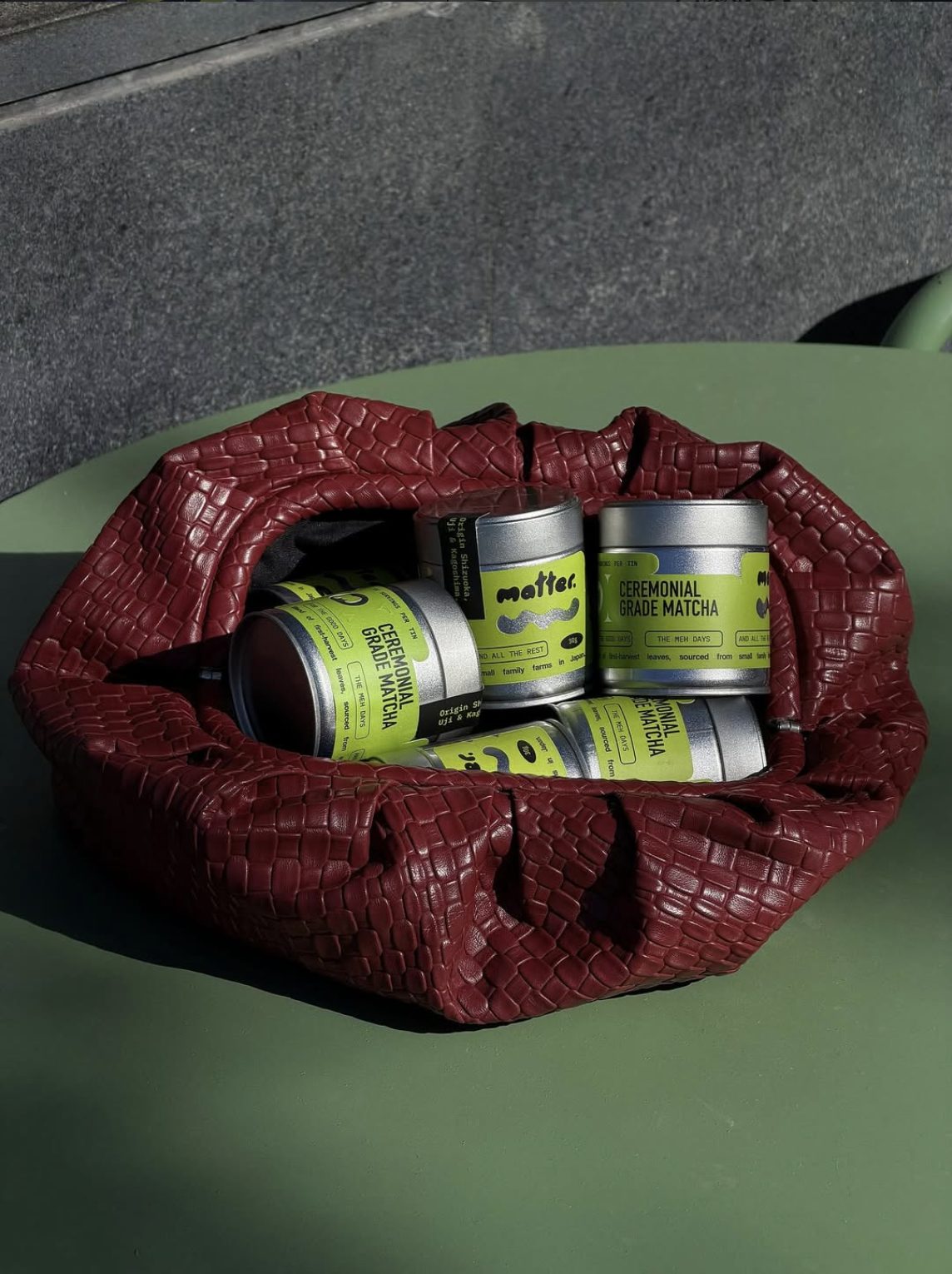 Silver tins of MATTER ceremonial grade matcha with acid-green labels, inside a burgundy woven leather bag on an olive table.